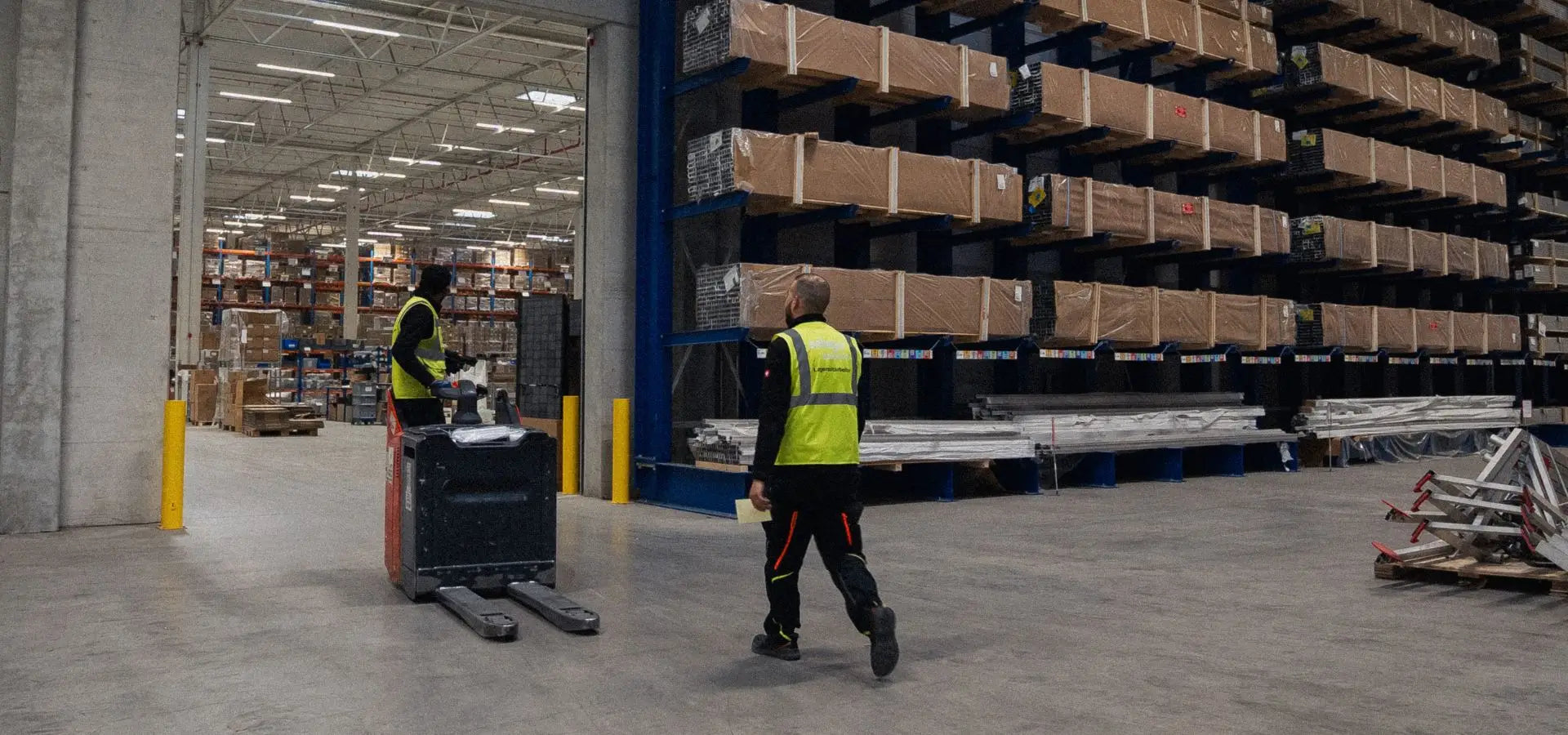 Zwei Lagermitarbeiter von solarhandel24 im Logistikzentrum, einer mit Elektro-Hubwagen vor einem Kragarmregal mit verpackten Solarmodulen und Aluminiumprofilen